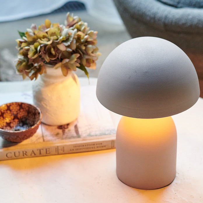 A small pale grey table lamp with a round base and a matching curved, solid dome shade next to a small vase of faux flowers.