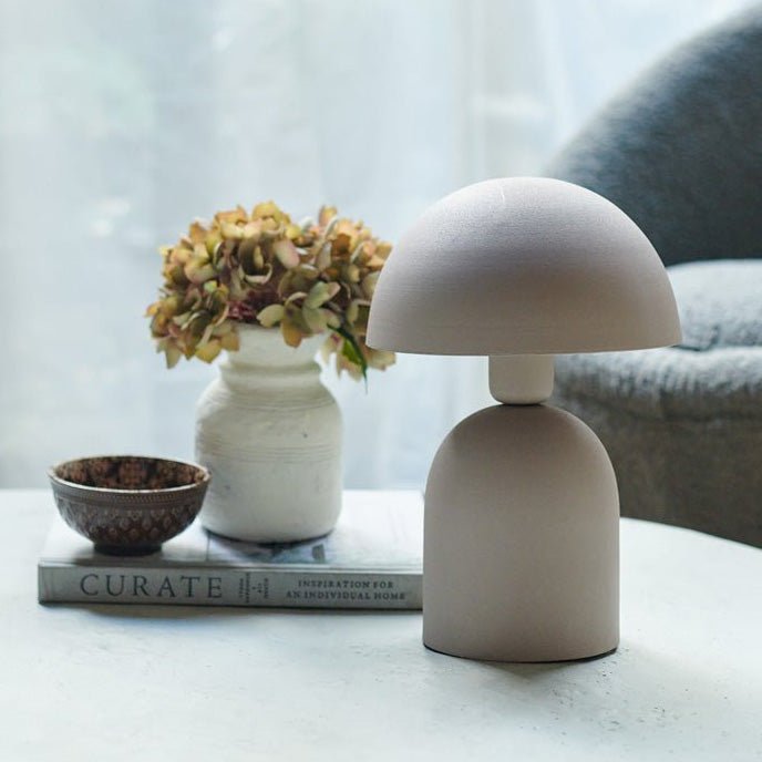 A small, soft grey table lamp with a round base and a matching curved, solid dome shade next to a small vase of faux flowers.
