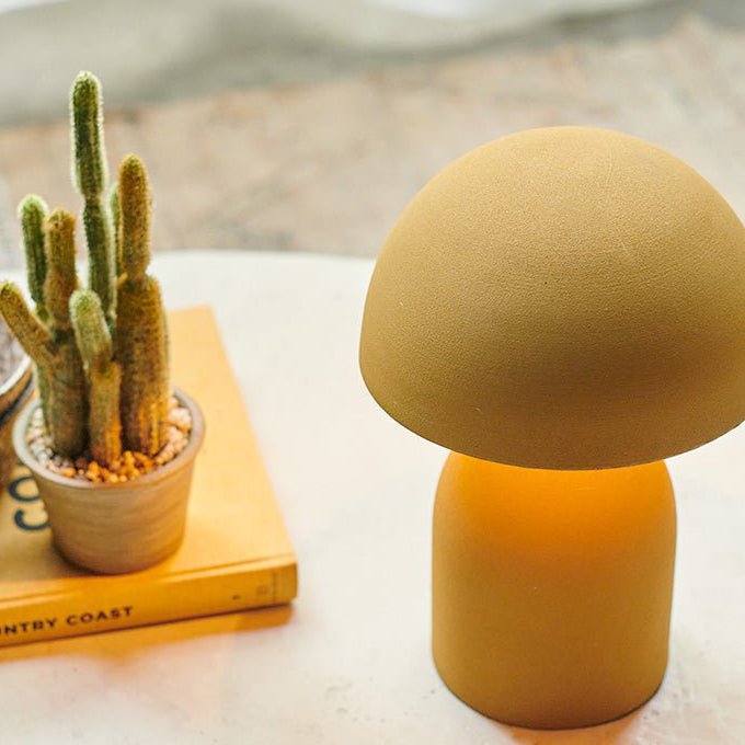 A small ochre table lamp with a round base and a matching curved, solid dome shade next to a cactus.