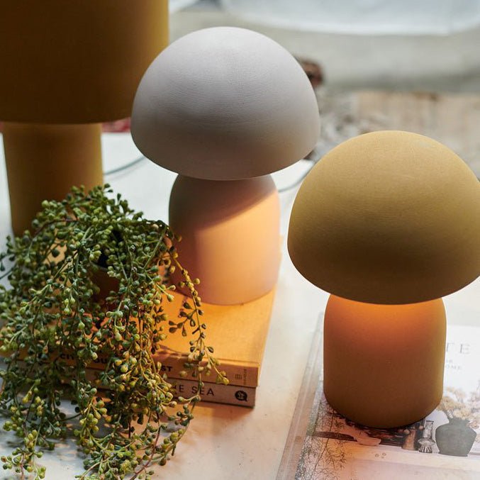 A small ochre table lamp with a round base and a matching curved, solid dome shade next to a matching lamp in pale grey and a small plant.
