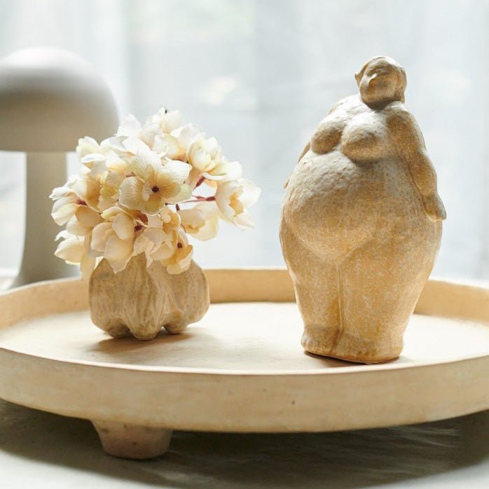 Ceramic sculpture of a standing female styled on a cream decorative tray next to a small vase of faux flowers.
