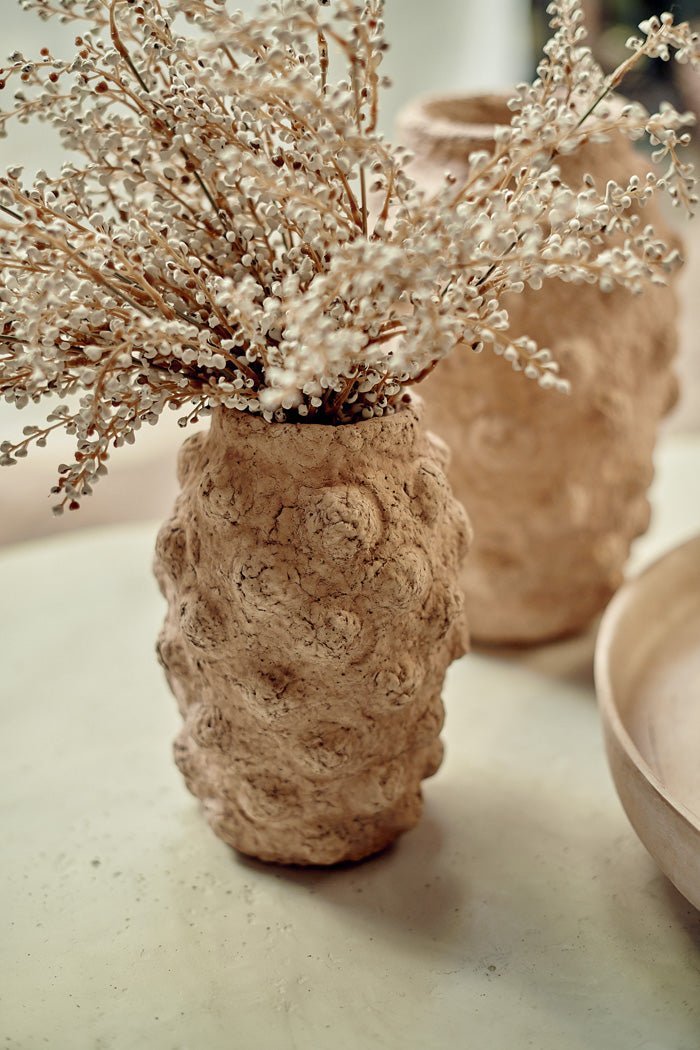 A textured beige cement vase with a bobble texture holding some artificial cream bud stems.
