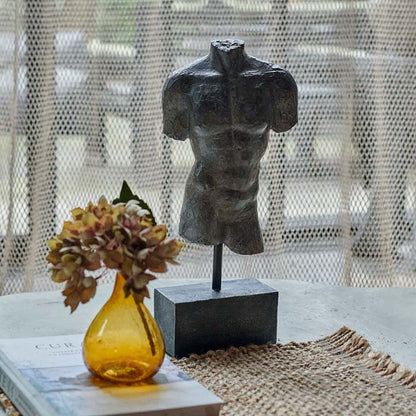 Image of the front of muscular torso in a classic figurative sculpture without any arms, on a solid block stand. The figurine is styled next to an amber vase with a premium faux flower.