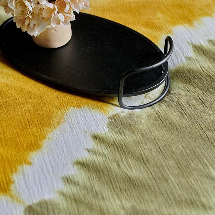 A closeup of the khaki and vibrant yellow tie-dye effect cotton fabric of the Saffron Bedspread, with an oval tray on it.