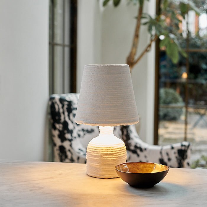 A small white paper mache table lamp creating a warm glow next to a ceramic bowl.