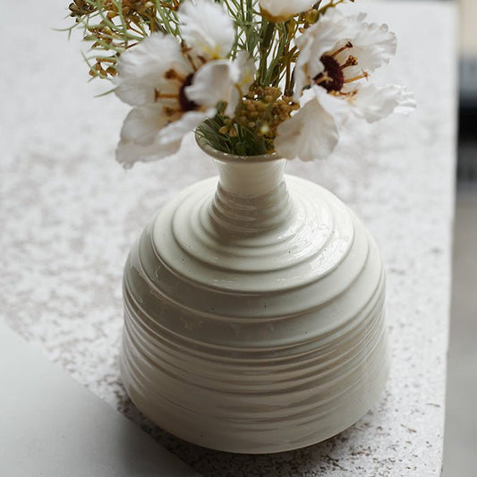 Styled image of a curved, ribbed white ceramic vase with a faux bouquet of flowers sitting on a countertop.