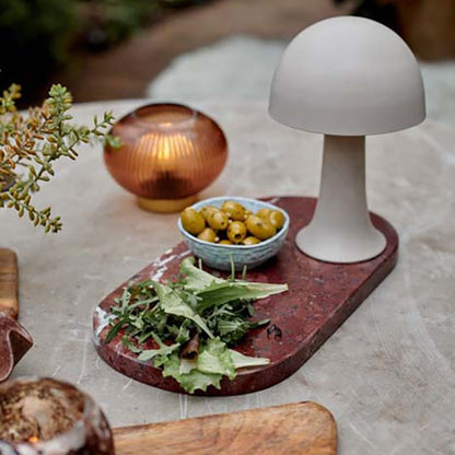 A small metal curvaceous mushroom shaped lamp in a soft grey colour by Abigail Ahern. This small battery operated lamp is sitting on a red marble serving board and is next to an amber glass LED lamp