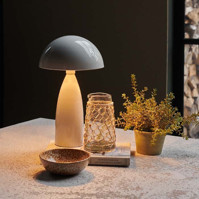 A pale grey table lamp with a dome shade and shiny finish giving off a warm glow next to some tableware and an artificial plant.