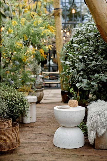 Styled image of a speckled, white curved side table in a garden amongst some foliage.