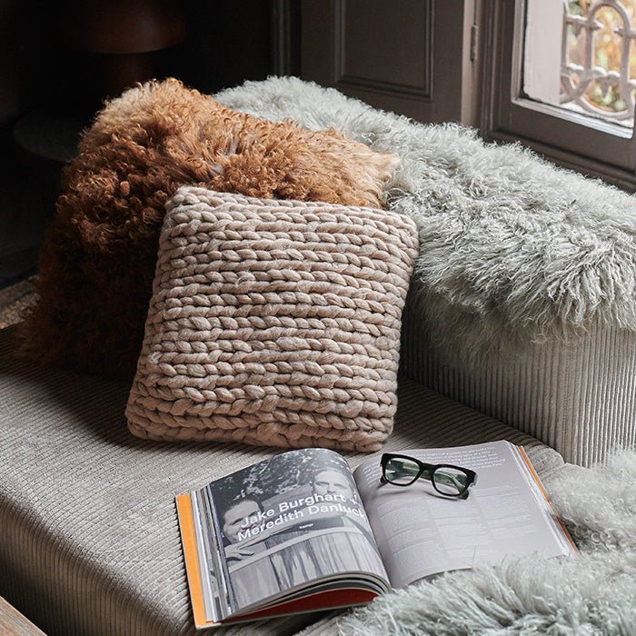 The Riva Cushion, a small square cushion in a soft natural blush shade with a chunky knit woven texture, next to an open book.
