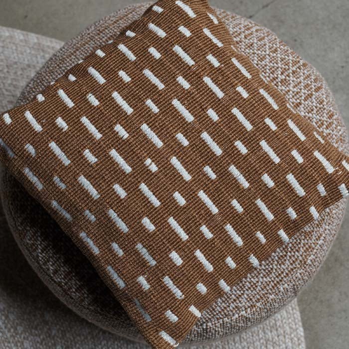 The Soriano Cushion, a square cushion with a woven cover in a rich earthy brown with white line detail, placed on a pouffe.