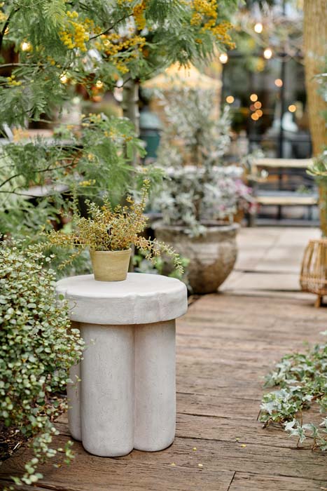 Styled image of a curved, smooth cream side table in a garden with a small fake plant on it.