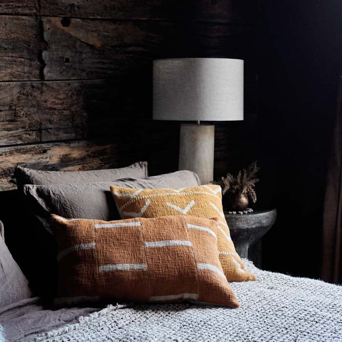 A rectangular cushion with a terracotta and cream cover with a simple brick design, sitting on a bed with a lamp in the background.