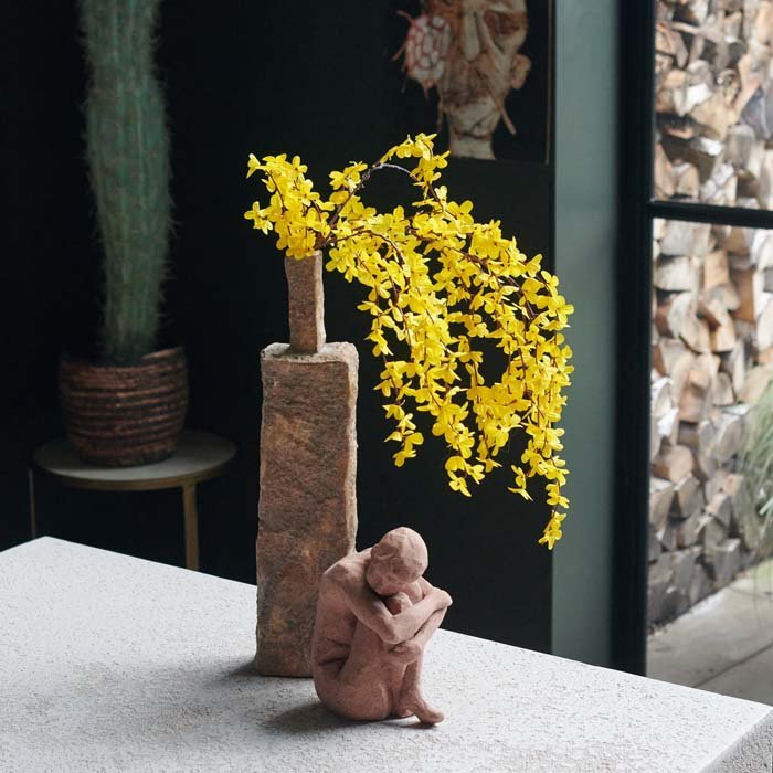 A small brown stoneware sculpture of a woman with her arms wrapped around her knees, in front of a terracotta vase with some yellow faux flowers.