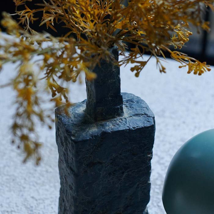 Detail of the rough texture on a dark grey porcelain vase in a rectangular style with some yellow artificial flowers.
