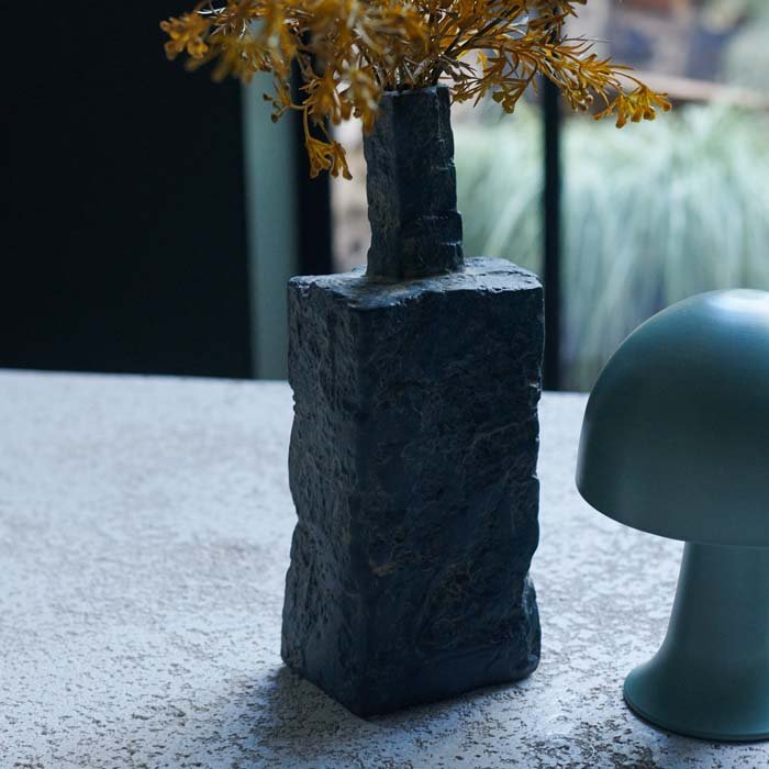 A rough textured dark grey porcelain vase in a rectangular style with some yellow artificial flowers and a small green dome table lamp.