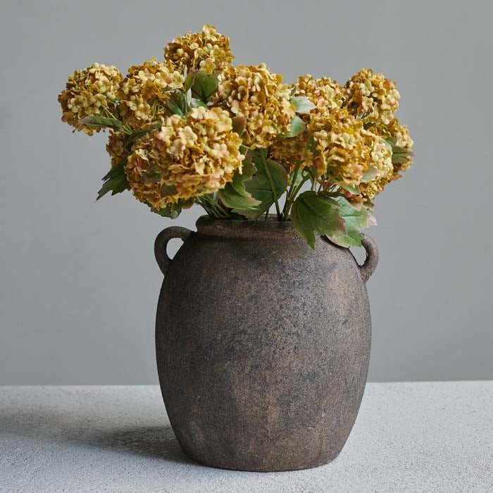 Isador Vase, a rounded ceramic vase in a rustic brown finish with two handles, containing some mustard coloured faux flowers.