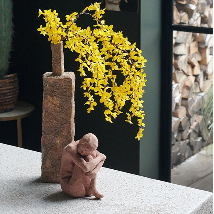 A small stoneware figurative sculpture in front of a terracotta vase with yellow artificial flowers.