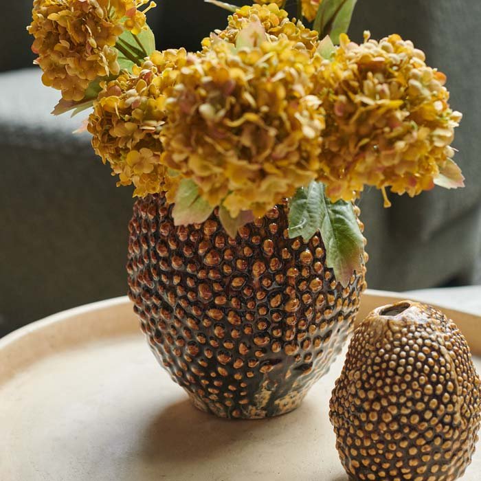 An organically shaped brown vase with a pebble texture holding a bouquet of yellow faux flowers