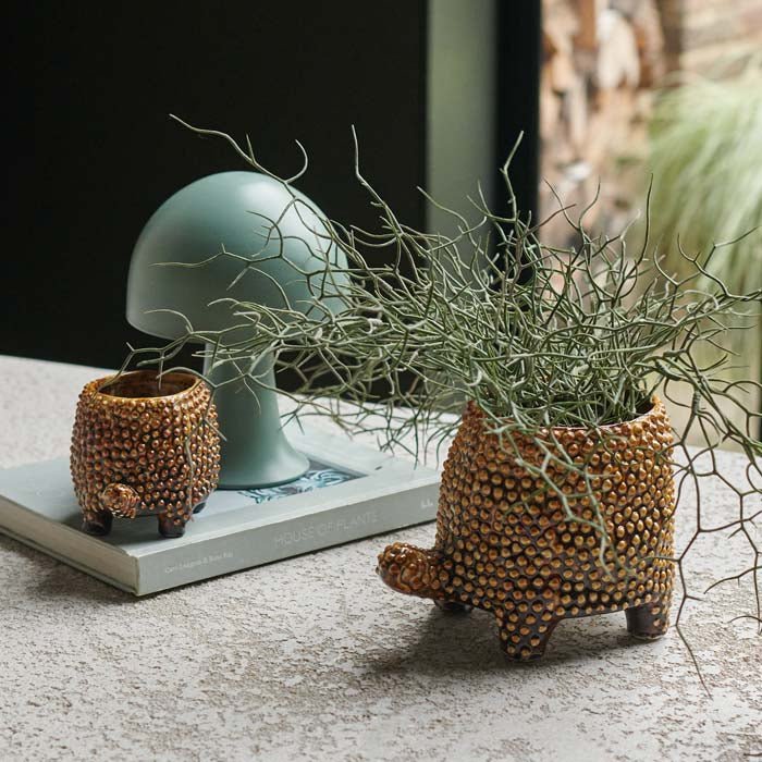 A brown tortoise shaped ceramic pot with a raised brown texture holding a faux plant on a table next to a smaller version and a lamp.