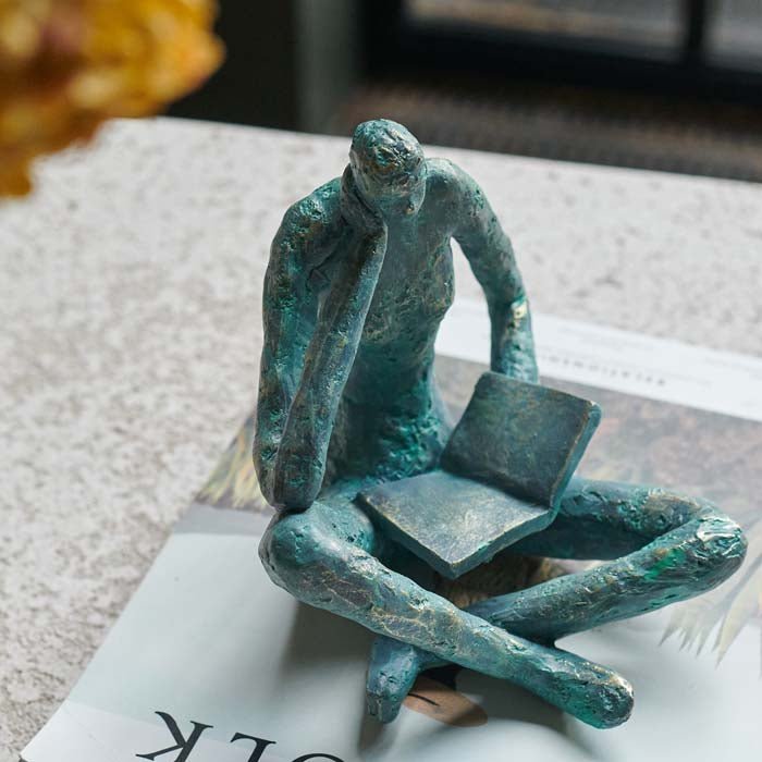 The Rollo Figurative Sculpture, a faux brass polyresin sculpture in a blue tone of a seated figure reading a book.