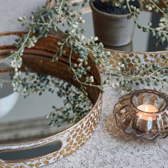 A closeup of the Vola Mirrored Tray Pair next to a votive and a beaded faux trailing potted plant.