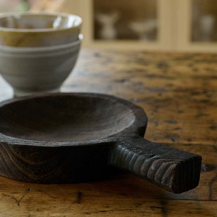 The Dagora Wooden Dish, a small round dish with a handle in a dark wood stain finish, next to a set of bowls inrustic hues.