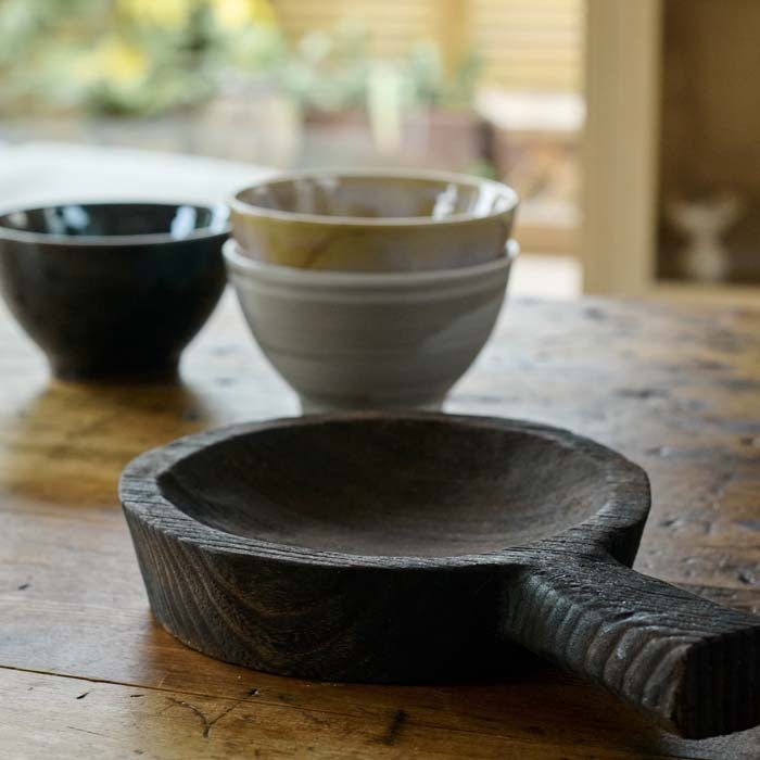 The Dagora Wooden Dish, a small round dish with a handle in a dark wood stain finish, next to a set of bowls inrustic hues.
