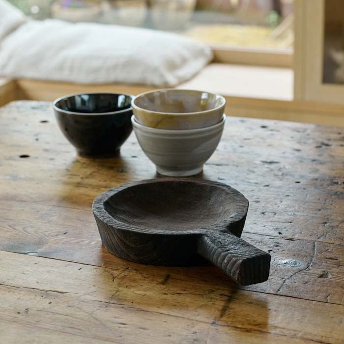 The Dagora Wooden Dish, a small round dish with a handle in a dark wood stain finish, next to a set of bowls inrustic hues.