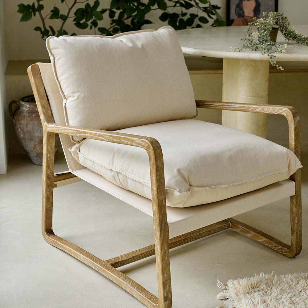 The Hockley Armchair, a modernist wooden framed armchair with cream cushioning next to a marble table and a tall plant.