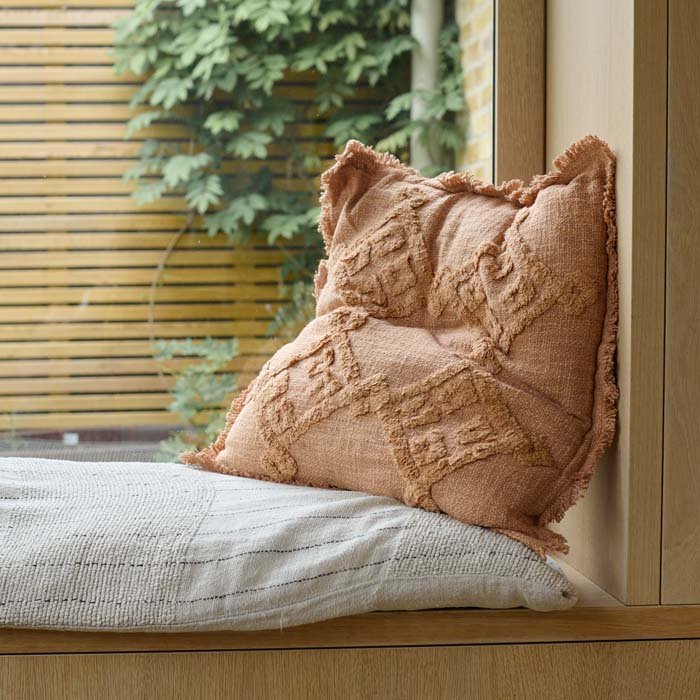 Pamra Cushion, a square cotton cushion in a soft rust colour with a tufted geometric pattern and a short fringe, by a window.