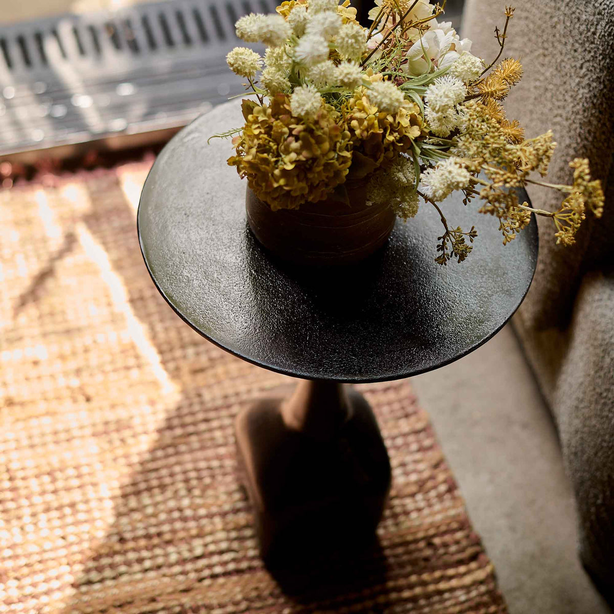 A small, round side table with a black metal finish holding a vase of faux flowers and sitting on a rug.