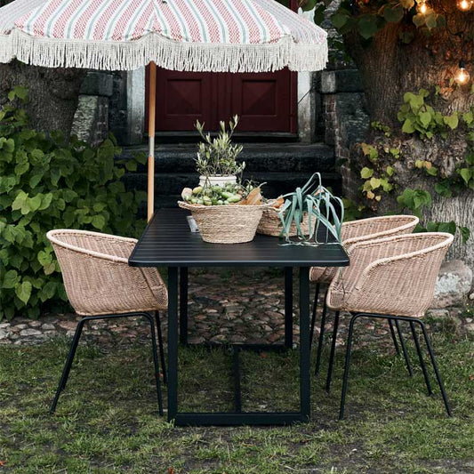 Lifestyle image of a black metal rectangular table outside with a garden umbrella, baskets and rattan chairs around it.