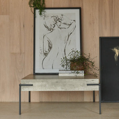An image of a faux concrete, rectangular table with four thin black metal legs, with a framed artwork of the sketch of a woman and some faux plants on top of it.