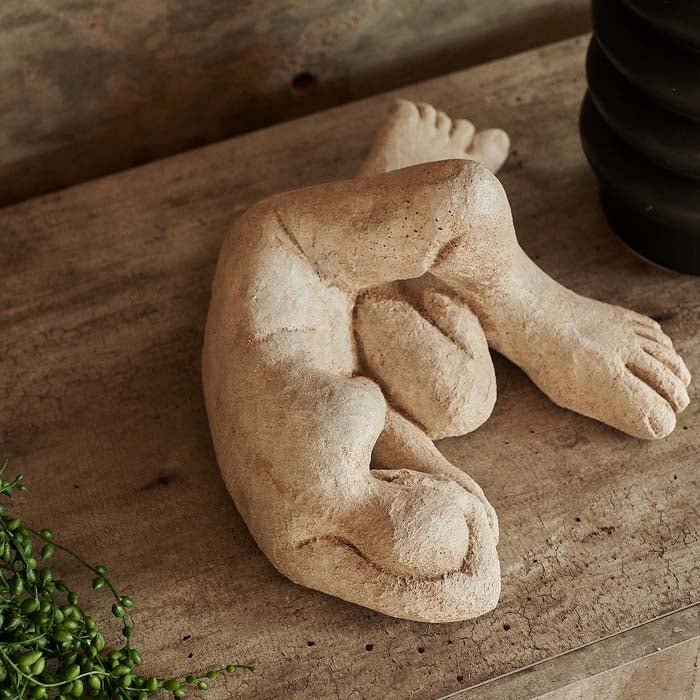 Cement figurative sculpture of a body lying on its side, with arms curled around its head and knees bent.