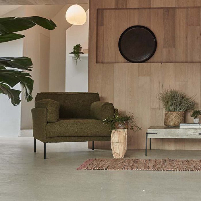 A green boucle fabric armchair with black iron legs, placed in a room with wooden flooring, a plant, and a decorative wall mirror.