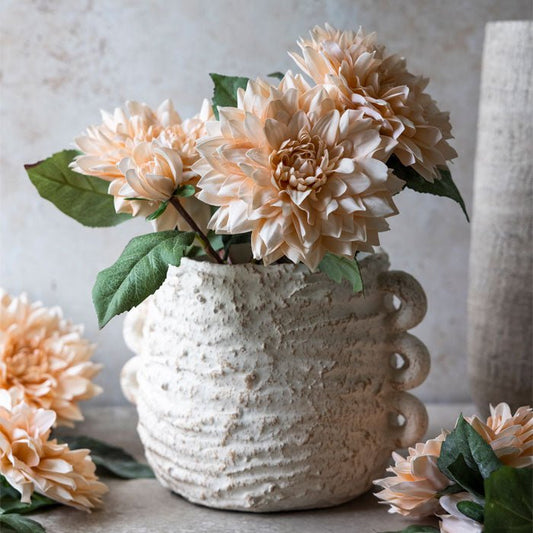Three peach coloured dahlia stems in a white textured vase surrounded by other dahlia stems