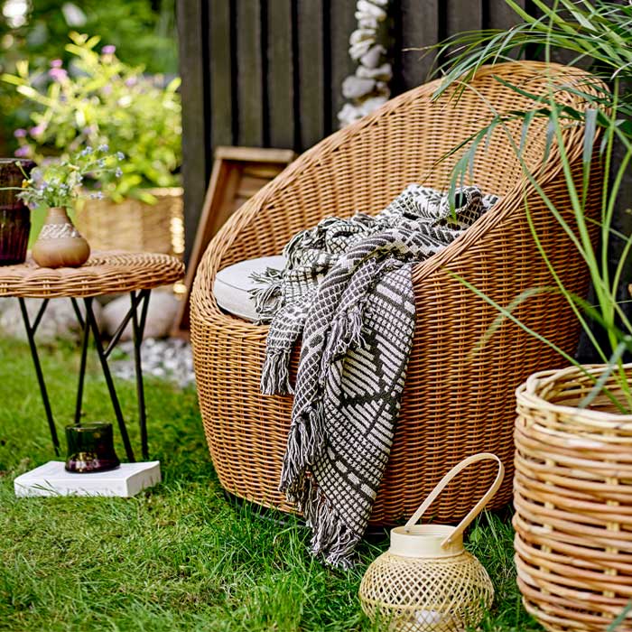 Lifestyle image of the woven bamboo lantern next to a woven lounge chair in a garden.