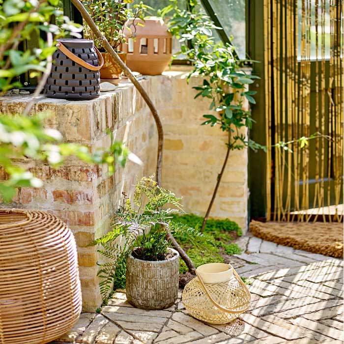 Lifestyle image of this woven hurricane lantern next to a brick wall with other lanterns and various plants.