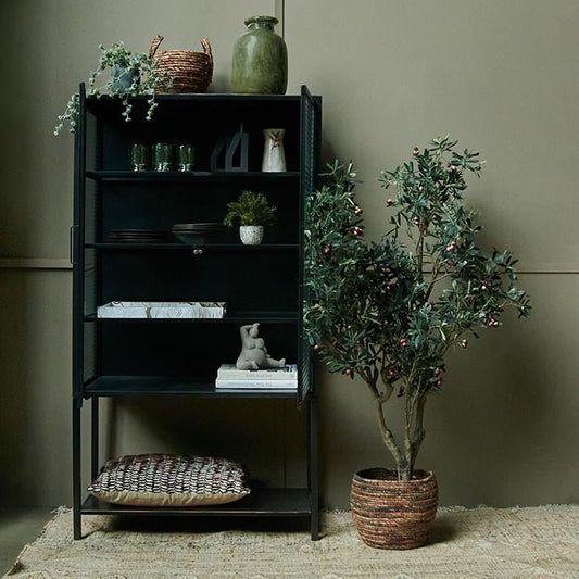 Large black iron cabinet open to show various home accessories and tableware inside, with an artificial olive tree next to it. 