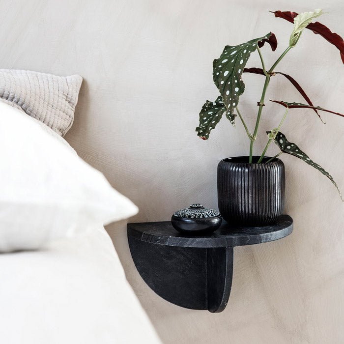 A black marble wall shelf made up of semi circle pieces with no visible nails. There's a plant pot placed on it and it's attached to the wall next to a bed.