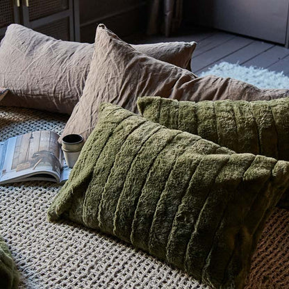 Image of two muted olive green cushions with some other soft furnishings. The pillows have a very soft, striped faux fur cover.