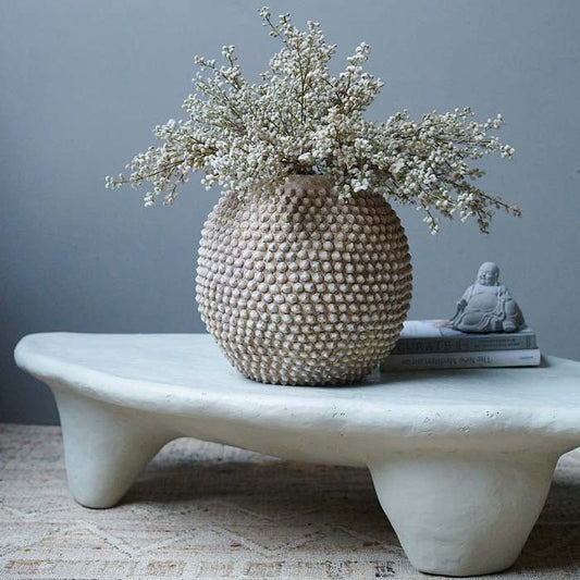 Pale brown, round vase with bobbled texture on the surface, styled with an artificial bundle and on a white coffee table.