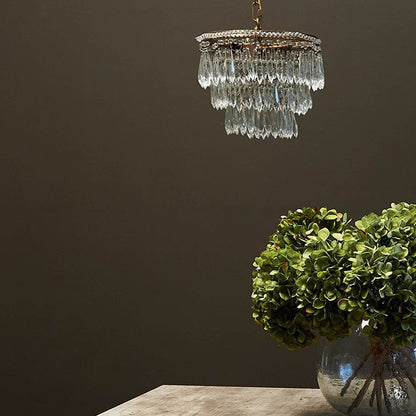 Styling image of a three tiered chandelier made of clear, teardrop glass beads on a brass chandelier frame, hanging about a bouquet of artificial flowers