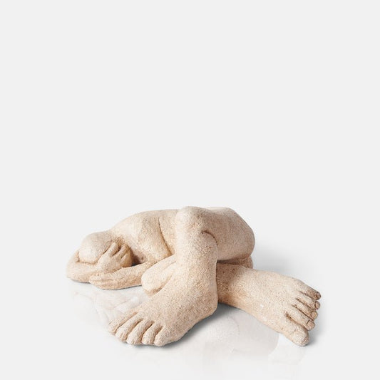 Cement sculpture of figure lying down with legs bent and arms curled around its head.