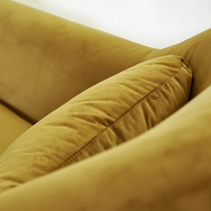 A closeup of the Atura Occasional Chair, a large angular yellow velvet armchair with a black metal frame and metal legs.