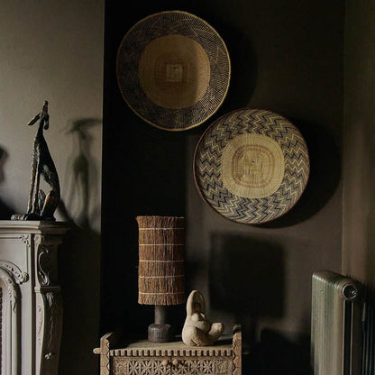 Two round patterned baskets hung on a brown wall above a table lamp and female sculpture