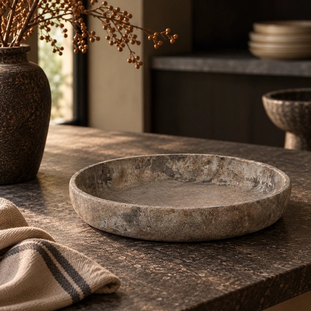 Broxtowe Cement Bowl with shallow rounded form and mottled stone-like surface, styled on a dark kitchen counter beside textured ceramics and neutral linens.