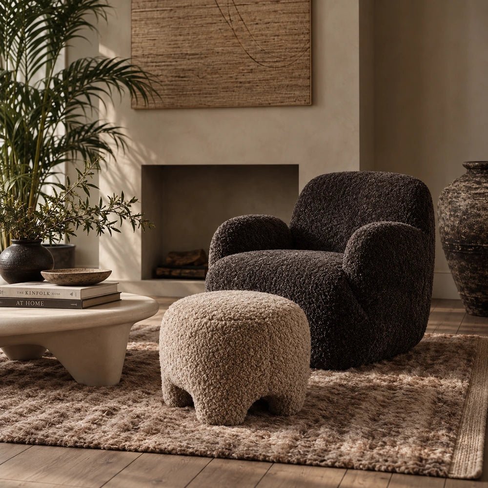 Calvello Faux Shearling Pouffe in a warm neutral living room, showing its soft cream textured upholstery beside a dark armchair and sculptural coffee table.