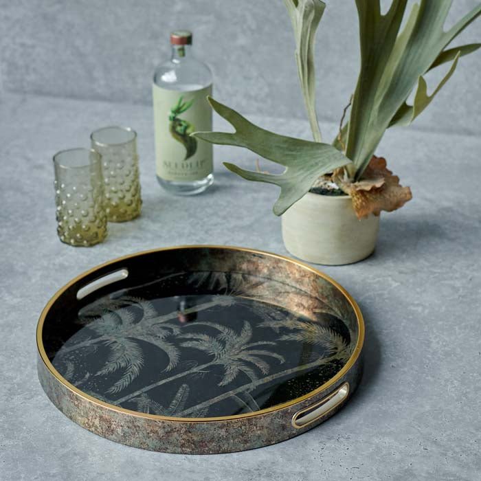 The Cayman Tray, a round tray with gold handles and a palm tree design in antique gold on black, next to a potted fern.